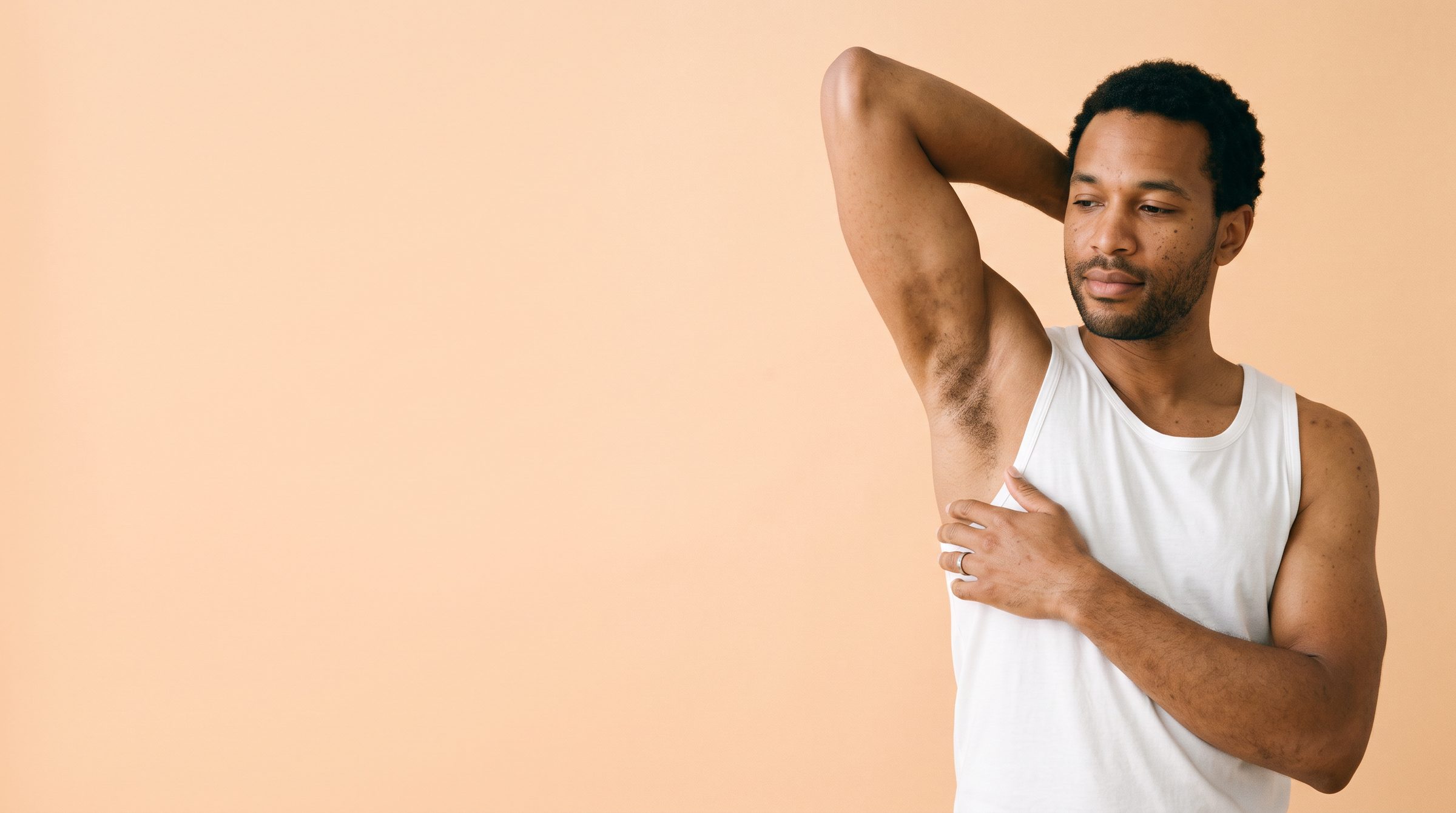 A man in a white tank top with one arm raised, showing the underarm